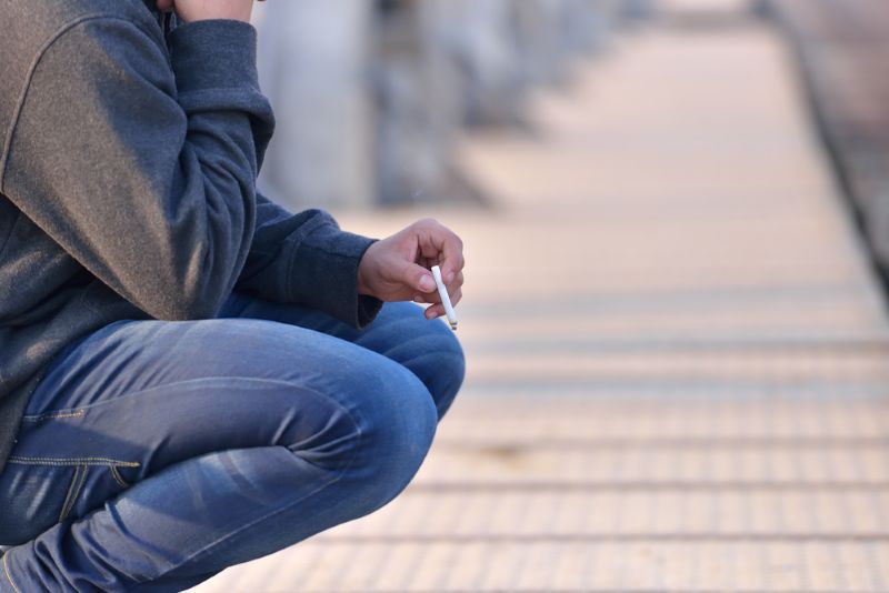Man wearing a sweatshirt crouched down while smoking a cigarette